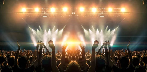Fotobehang Foto van een concertzaal met silhouetten van mensen die klappen voor een groot podium verlicht door schijnwerpers. De opname is gemaakt vanuit het oogpunt van de concertmenigte, lensflare is zichtbaar. © CesareFerrari