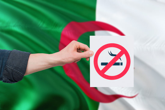 Algeria health concept. Hand holding paper with no smoking sign over national waving flag. Quit smoke theme.