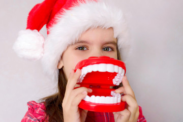 happy funny baby girl in red Christmas hat with toy model teeth on grey background.