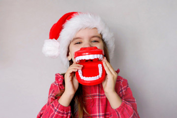 happy funny baby girl in red Christmas hat with toy model teeth on grey background.