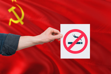 Soviet Union health concept. Hand holding paper with no smoking sign over national waving flag. Quit smoke theme.