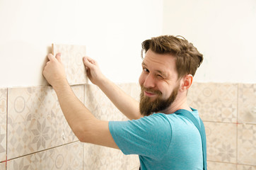 Charismatic handyman winking while putting on tiles on the wall 