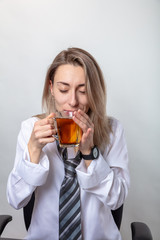 A blond woman in a white shirt and tie is drinking tea. There is a break in the train.
