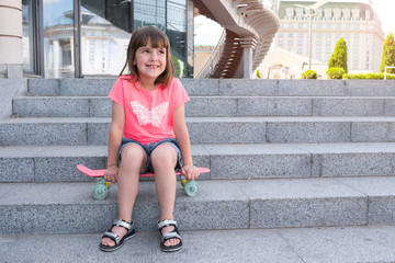 Fashionable little girl is sitting on a skateboard in the city. Life style.