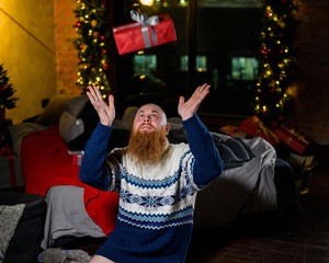 A man with a long red beard sits on the floor without pants in a winter sweater. man is holding a wrapped present on the background of New Year decorations and lights. Christmas tree. Parody, humor.