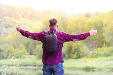 a man with a backpack, with arms spreading to the sides, stands by the river and looks at the bright sunny sky. concept of travel and freedom