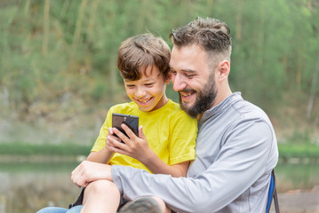 man and a boy are sitting on a background of green trees and smiling are looking at a mobile phone. concept of family paternity