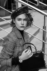 Outdoor monochrome portrait of elegant woman wearing trendy beret, checkered coat, stylish wrist watch, holding black leather classic handbag,  posing in street