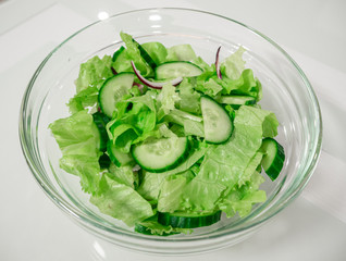 transparent bowl with green salad, cucumbers, red onions on a white table or background, top view