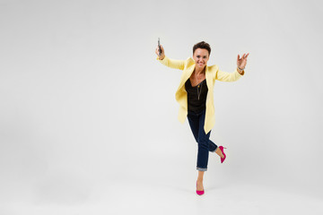 Fototapeta na wymiar pretty brunette with a short haircut in a cream-colored jacket and pink pumps pumps dancing with a phone in her hands on a gray background