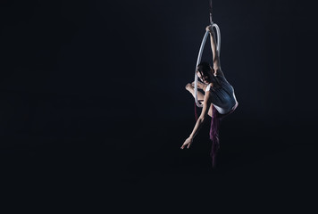 Young woman performing acrobatic element on aerial ring against dark background. Space for text