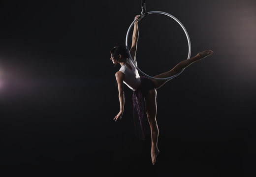 Young woman performing acrobatic element on aerial ring against dark background
