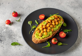 rye bread toast with mashed avocado, roasted chickpeas, herbs on a dark plate. clean eating, dieting. gray concrete background, side view