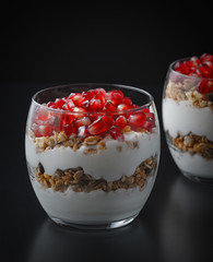 Yogurt with granola and pomegranate fruits in a glass cup on a black wooden table. Tasty dessert Healthy breakfast