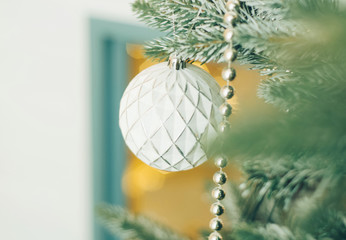 White Christmas ball toy and a silver beads on the snow branch of a green tree. Festive yellow lights from house window