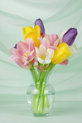 close up of colorful tulips floral arrangement in a clear vase on an elegant draped light green fabric background