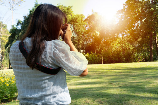 Woman standing and taking photos on the lawn facing the forest with sunlight. Women wear comfortable outfits when traveling.