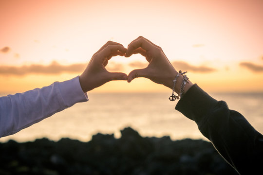 Silhouette image of two young female hands making heart on a beautiful summer evening. Girl fingers like hearts sunset orange sky  and sea in background Enjoyable romance feelings relationship concep