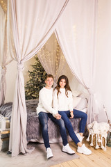 Obraz premium Young beautiful couple sit on the bed on the background of the Christmas tree and smiling