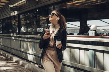Portrait of Stylish Girl Wears Women Overalls and Black Leather Jacket Drink Coffee, Using Smartphone