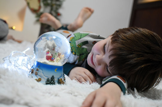 Boy looking a glass snow ball under a Christmas tree. Child with a snow ball with a New Year - Powered by Adobe