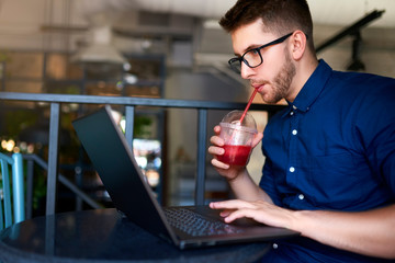 Freelancer man with plastic cup of fresh beverage in hand works with laptop. Businessman in glasses drinks juice for body hydration while working. Attractive designer quench thirst. Healthy lifestyle.