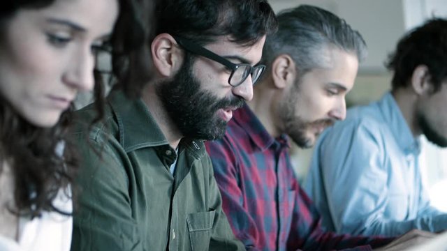 Side view of smiling developer in eyeglasses working with laptop. Young people sitting in row and typing on laptops. Technology concept