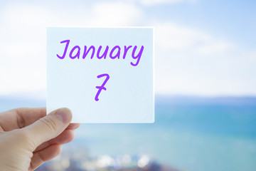January 7th. Christmas day. Hand holding sticker with text January 7 on the blurred background of the sea and sky. Copy space for text. Month in calendar concept
