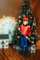 6 years old boy wearing like hussar with saber stay near christmas tree. Big clock and dollhouse on floor. Child in masquerade carnival costume of nutcracker. Dreams come true in christmas time