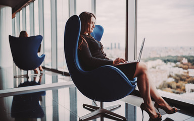 A charming caucasian business lady is sitting with a laptop on an armchair of classic blue color in front of a huge panoramic window, on the top floor of a luxury office skyscraper and looking outside
