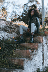 Smiling African female in a leather jacket is sitting on antique stony steps outdoors; a vertical shot of a cheerful black Guinean girl with a curly hair is sitting on an old flaked outdoor stair