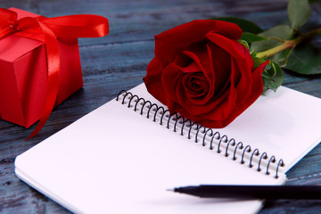 Valentine's day background. On a blue wooden background an opened notebook, pen, gift and red rose. Close-up, horizontal, side view, free space for text.
