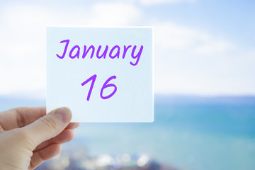 January 16th. Hand holding sticker with text January 16 on the blurred background of the sea and sky. Copy space for text. Month in calendar concept