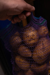 Mesh bag with potatoes on a dark background. Potato packaging. A man's hand holds a net with vegetables.