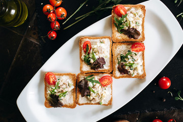 Brushetta set. Variety of small sandwiches with egg, tuna, arugula and salad served with glass of cognac on a black stone board over dark background