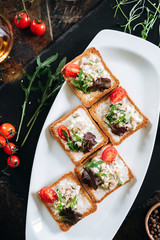 Brushetta set. Variety of small sandwiches with egg, tuna, arugula and salad served with glass of cognac on a black stone board over dark background