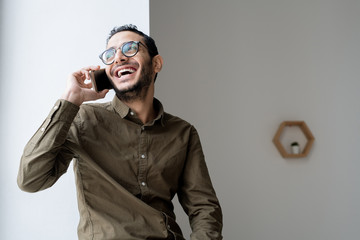 Happy young casual businessman talking on mobile phone by office window