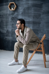 Young pensive businessman in smart casual sitting on chair in front of camera