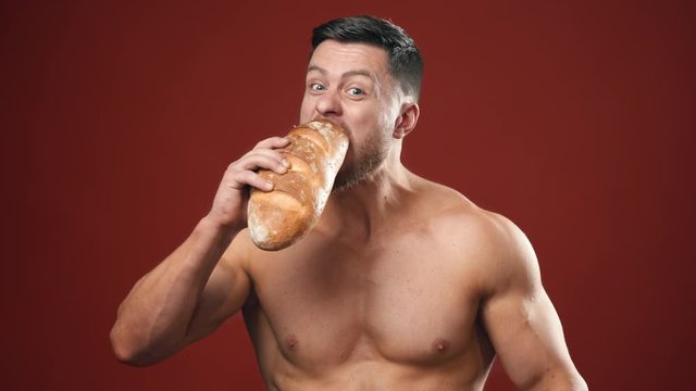 Hungry strong male eating unhealthy food. Half-length portrait of muscular man without shirt eats bread and sausage quickly on the studio background.