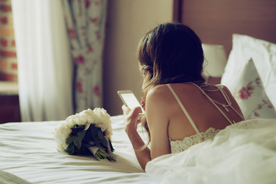 Morning of the bride. Beautiful European bride in gorgeous white gown using mobile phone.