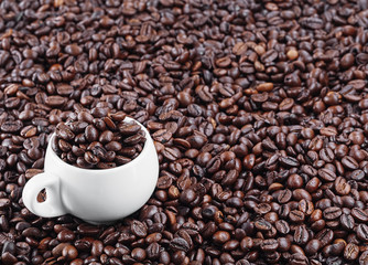 Roasted coffee beans, background. Horizontal orientation. Fresh aromatic dark coffee. A white cup for espresso stands in coffee beans. Close-up. Selective focus on a cup with coffee grains.