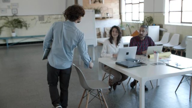 Back view of curly man walking to cheerful colleagues. Group of smiling young people working together in cozy office. Startup concept