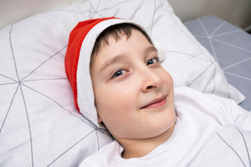 A preteen boy lying in bed in santa hat during christmas eve night and waiting for gifts and miracles, happy xmas and new year holiday