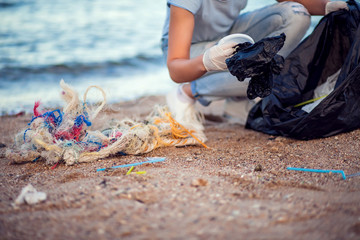 Woman with white gloves and big black package collecting garbage on the beach. Environmental protection and planet pollution concept