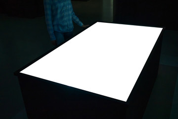 Woman looking at white blank interactive touchscreen display of electronic multimedia table kiosk in dark room of modern technology exhibition. Mock up, copyspace, template and futuristic concept