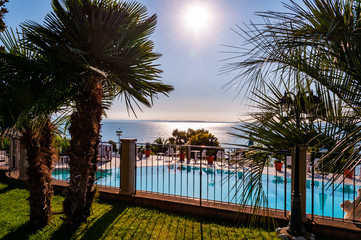 Sunny outdoor pool behind the fence with crystal water, parasols and deck chairs with Garda lake and Sirmione city on the background