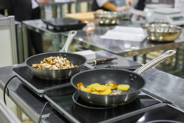 Process of cooking sliced champignon mushrooms and potatoes in frying pans with oil on electric stove at cuisine of restaurant. Professional cooking, catering, cookery, gastronomy and food concept