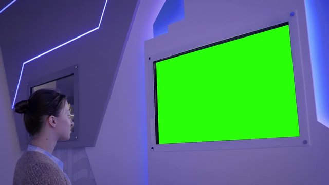 Green screen, mock up, future, copyspace, template, chroma key, technology concept. Woman looking at blank digital interactive green display wall at exhibition or museum with futuristic scifi interior