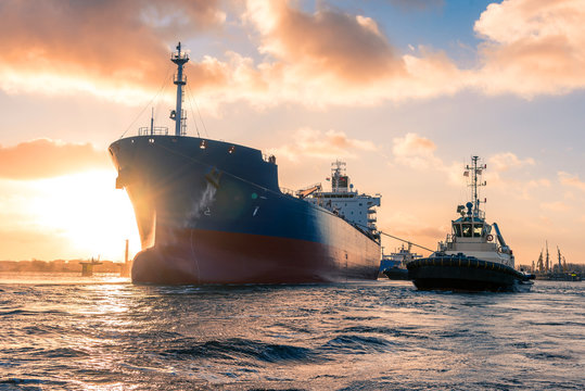 Amazing cloudy sunrise during mooring operations in port. The huge tanker is receiving tugboat assistance to navigate safely to the jetty. Merchant, maritime action background filled with sun rays.