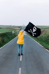 A young girl with a yellow raincoat walks along a road between fields heading to the coast waving a pirate flag.